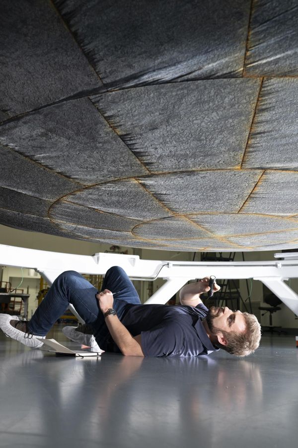 An engineer lying on a mat on the floor looks up at the underside of the Orion heat shield, its dark blocks separated by orange-tinted seams
