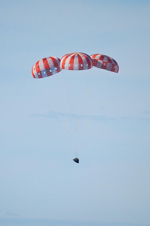 Orion hanging beneath three large orange-and-white parachutes against a pale blue sky