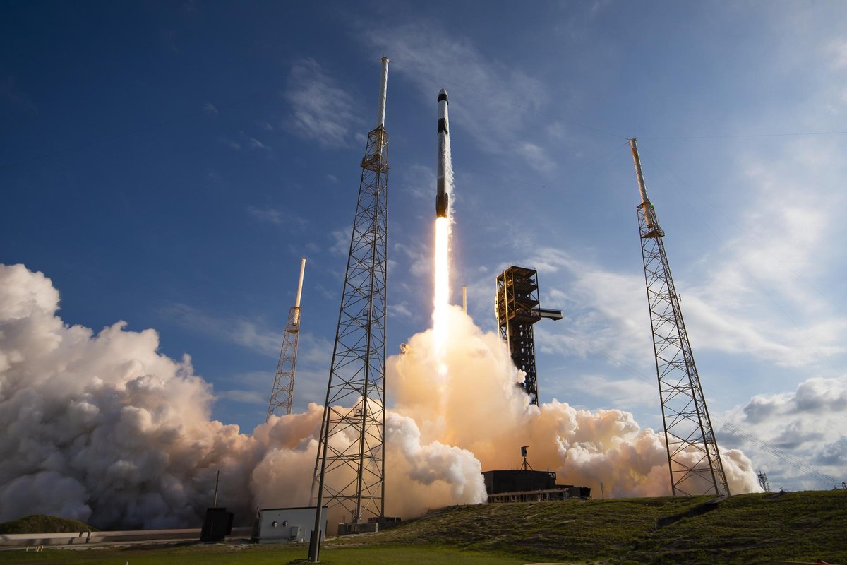 A SpaceX Falcon 9 rocket lifts off from a coastal launch pad in a burst of white exhaust under blue sky