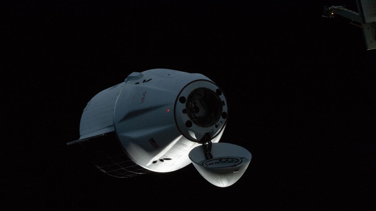 A white SpaceX cargo Dragon with its nose cone open approaches the space station against black space