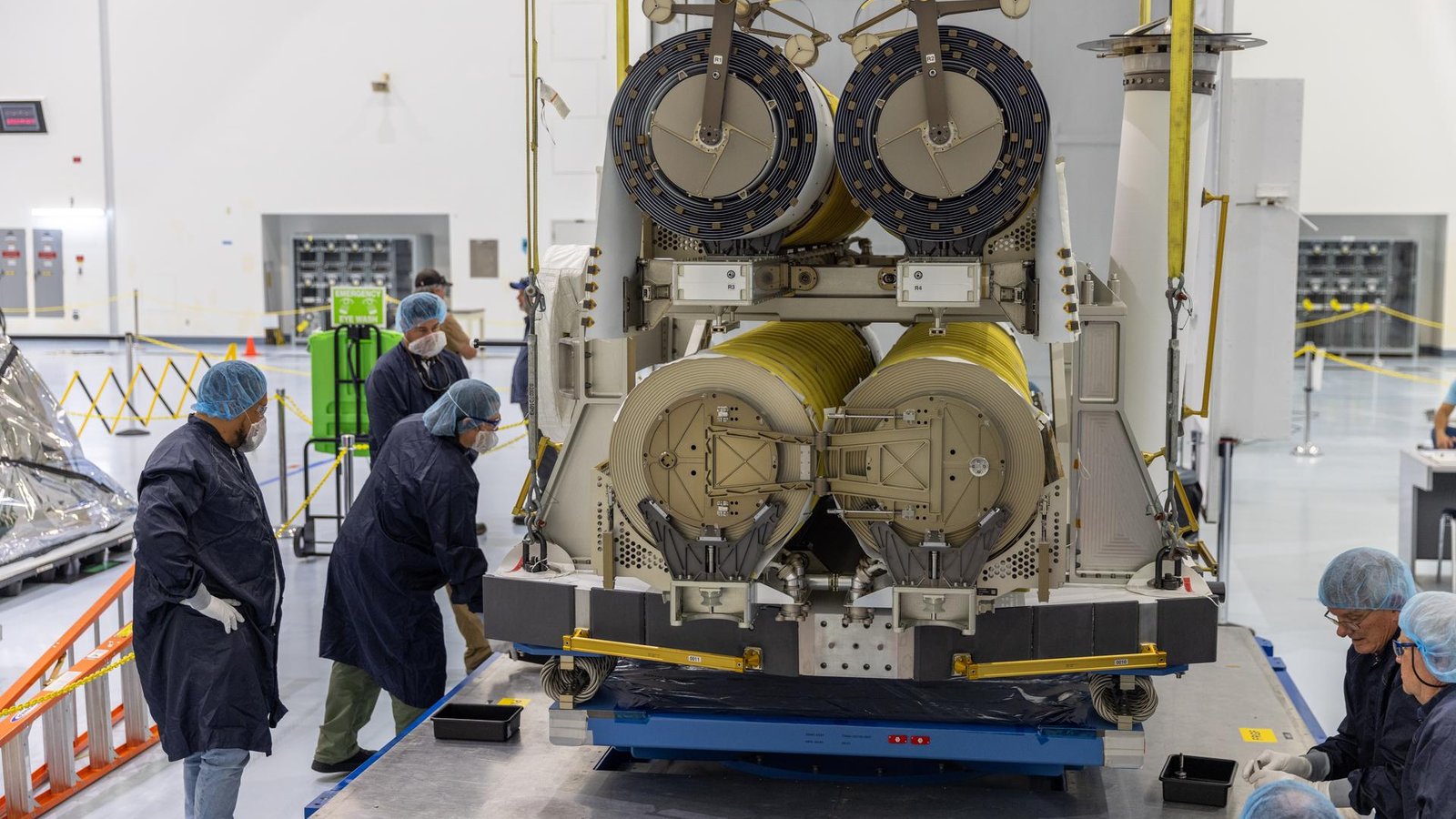 Technicians in protective gowns guide a crane as it lowers a pair of rolled-up International Space Station Roll-Out Solar Arrays onto a platform inside NASA Kennedy’s Space Systems Processing Facility