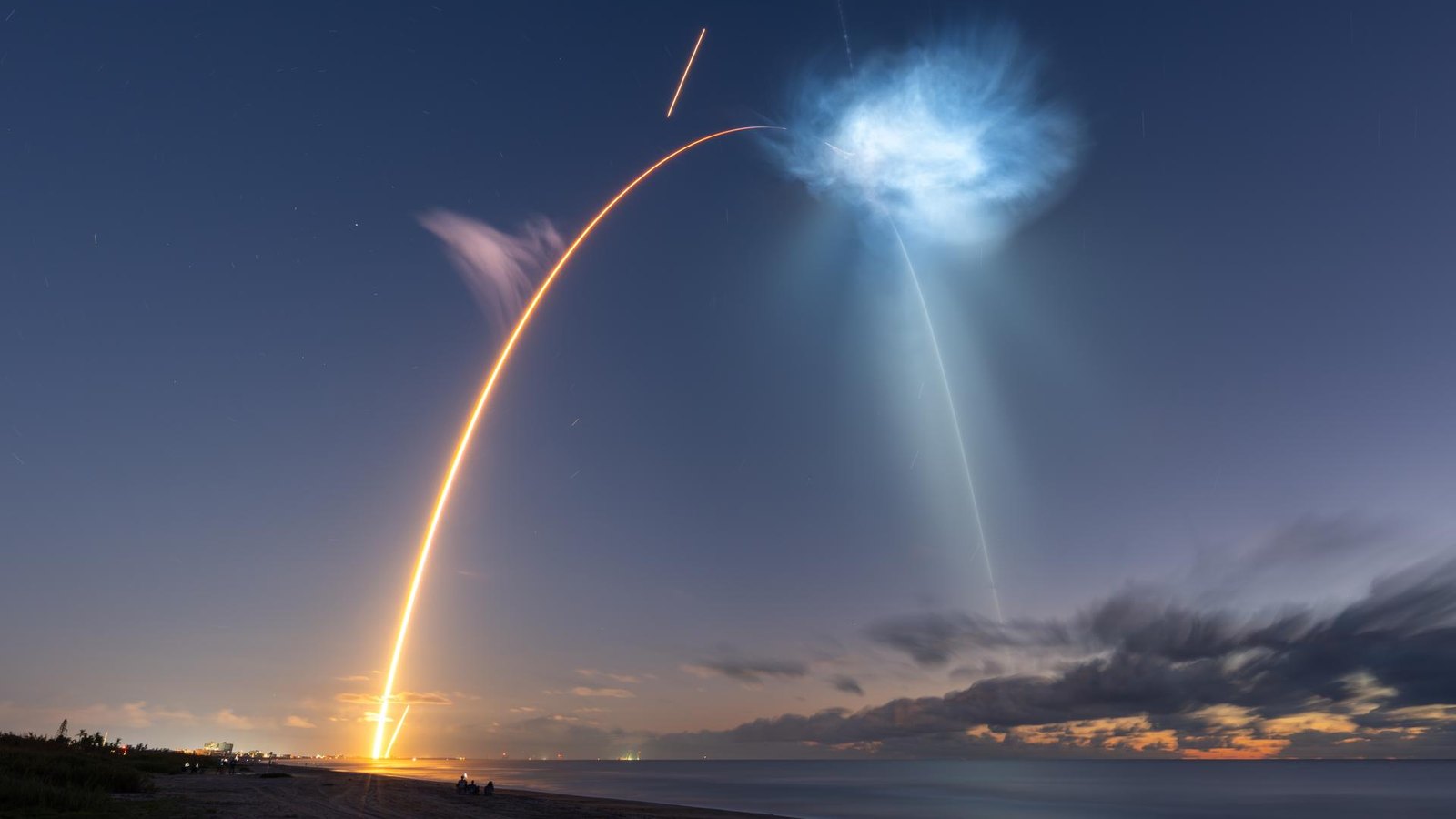 Long-exposure photo from a beach at twilight: a Falcon 9 launch streak arcs up from the horizon beneath a glowing exhaust cloud
