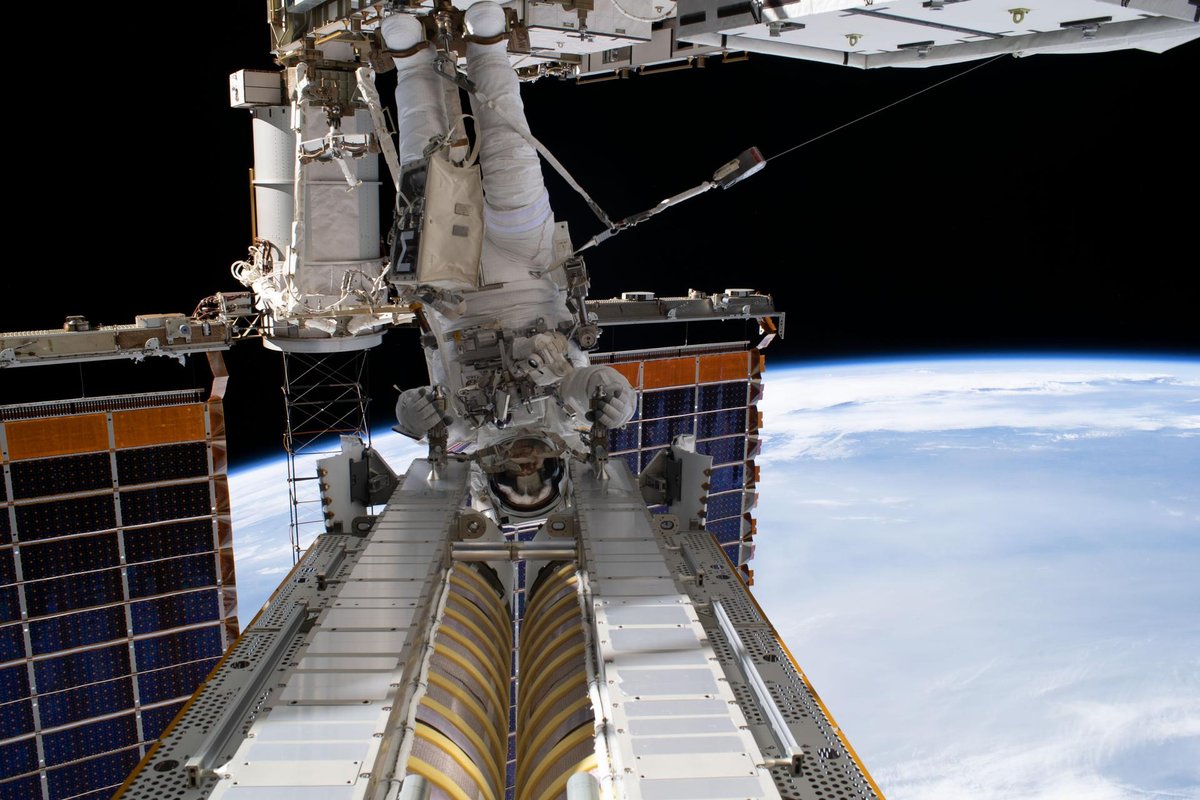 An astronaut in a white spacesuit, attached to the station’s robotic arm, handles a rolled-up solar array above Earth during a spacewalk