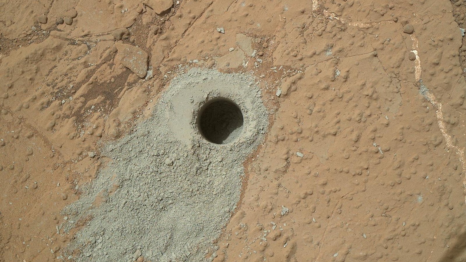 Close-up photograph of the Cumberland drill hole in Gale Crater mudstone: a dark circular hole surrounded by gray powdered rock on reddish Martian bedrock