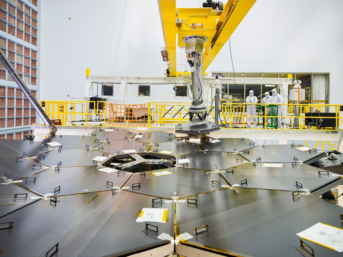 Technicians in white clean-room suits watch from a yellow railing as a robotic arm sets the last gold-coated hexagonal segment into the James Webb Space Telescope’s primary mirror