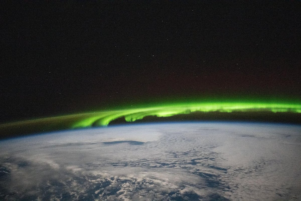 A bright green aurora band arcing along Earth’s horizon above a cloudy ocean, photographed from the International Space Station at night