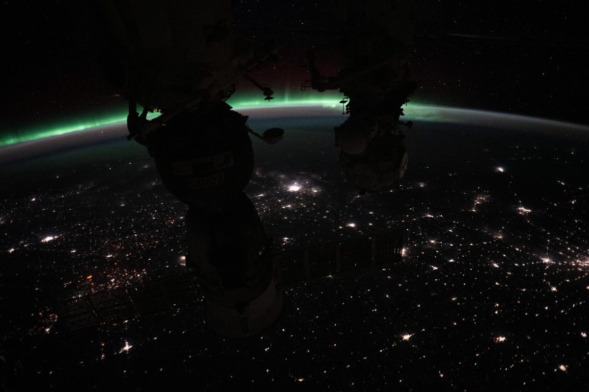 Thin green aurora arcing along Earth’s horizon above Canada at night, with city lights below and dark space station hardware silhouetted in the foreground