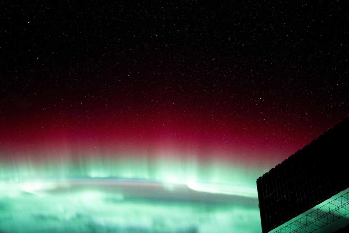 Red and green aurora borealis glowing above Earth’s horizon, photographed from the International Space Station, with a station solar array at lower right